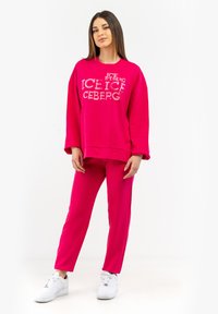 Fuchsia sweatshirt with “ICEBERG” print, paired with matching fuchsia pants. Both made from soft fabric, complemented by white sneakers.