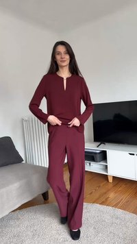Kobieta poprawiająca bordowy top z długim rękawem i dopasowane szerokie spodnie, stojąca na okrągłym dywanie w salonie z telewizorem i sofą.