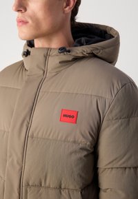 Olivovo zelená prešívaná bunda so zipsom vpredu, kapucňou na stiahnutie a viditeľnými sekciami; obsahuje červený obdĺžnikový štítok s logom "HUGO".