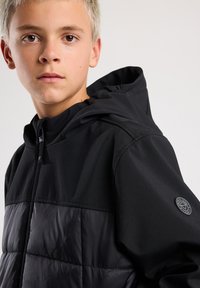 Musta takki, jossa on sileä yläosa ja kiiltävä pehmustettu alaosa, huppu sekä pyöreä logo-patch hihassa.