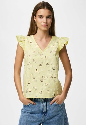 Frau steht mit den Händen in den Taschen, trägt ein gelbes geblümtes Lochspitzentop mit gerafften Ärmeln und blaue Jeans.