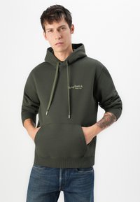 Young man wearing dark green hoodie with pockets, blue jeans, and showing tattooed left forearm, standing against white background.