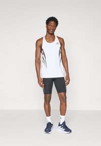 Vit träningslinne med svarta detaljer, tillsammans med svarta shorts. Har en rund halsringning och Adidas-logotyp. Modellen bär sneakers.