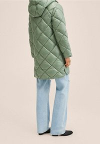 Abrigo acolchado verde con capucha, con patrón de rombos, llevado sobre unos vaqueros de pierna recta de color azul claro. Textura suave, diseño hasta la rodilla.