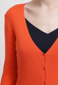 Cardigan lavorato a maglia arancione con scollo a V, dotato di chiusura con bottoni e maniche lunghe. Texture morbida con dettagli a coste sul colletto.