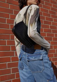 Svart krokodilpräglad väska med en slät yta, bärs över axeln, i kombination med blå denimshorts och en långärmad mönstrad topp.
