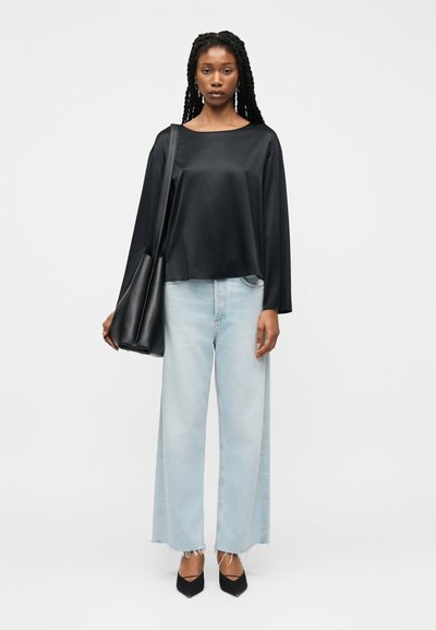 Femme aux cheveux tressés portant un chemisier en satin noir ample, un jean large clair avec des ourlets effilochés, des talons noirs, et portant un grand sac noir à l'épaule.