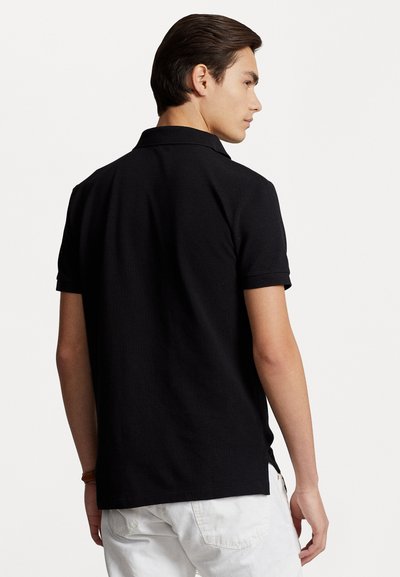 Svart polo-skjorte med korte ermer, kragehals, teksturert stoff og ren silhuett, brukt med hvite shorts, vist fra baksiden.