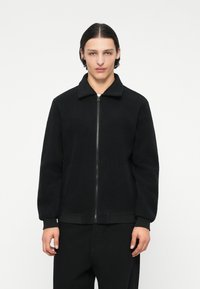 Giacca in pile nera con zip frontale, colletto e polsini a costine. Presenta una superficie testurizzata e una vestibilità rilassata. Perfetta per un look casual.