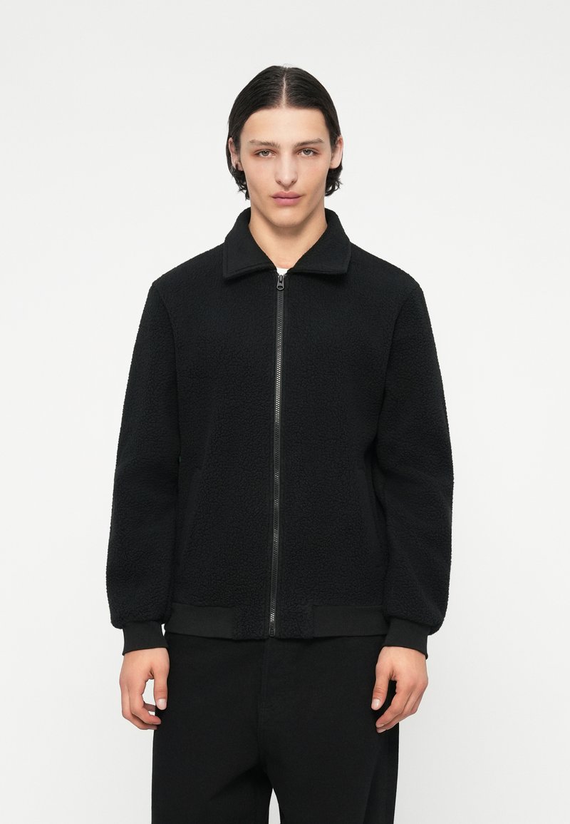 Giacca in pile nera con zip frontale, colletto e polsini a costine. Presenta una superficie testurizzata e una vestibilità rilassata. Perfetta per un look casual.