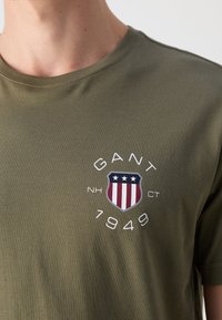 Olivgrön bomulls-t-shirt med rund halsringning, korta ärmar och en sköldlogotyp med röda, vita och blå accenter samt texten "GANT 1949".