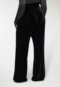 Osoba ubrana w czarne, szerokie, welurowe spodnie oraz długą, ciemnoszaro top, pokazana od tyłu na jednolitym tle.