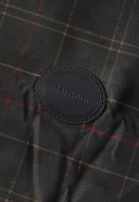 Stofa tartan întunecată cu linii roșii și galbene, având o pată ovală neagră cusută, embosată cu numele mărcii "Barbour".