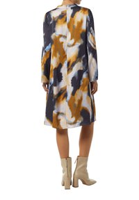 Langärmliges Kleid mit einem mehrfarbigen abstrakten Muster in Marineblau, Gold und Hellblau; hat einen geraden Schnitt und einen Rückenschluss. Kombiniert mit beigen Knöchelstiefeln.