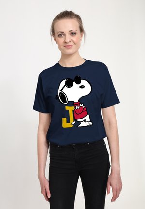 Donna che indossa una maglietta blu navy con una grafica di Snoopy con occhiali da sole e camicia rossa, in piedi accanto a una grande lettera J gialla.