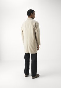 Beige kabát laza szabással, egyenes aljjal és hegyes gallérral, sötétkék nadrággal és barna cipővel párosítva.