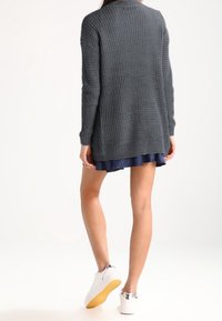 Grå stickad cardigan med långa ärmar, texturerat tyg och avslappnad passform, matchad med en marinblå volangkjol och vita sneakers.