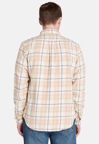 Camisa a cuadros beige con mangas largas, puños abotonados y detalle de pliegue en la parte posterior. La tela tiene una textura suave con patrones de cuadros claros y oscuros.