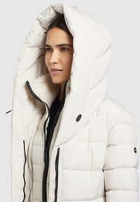 Puffjacke in hellbeige mit großer Kapuze, ausgestattet mit einer gesteppten Textur, Reißverschluss und schwarzen Akzenten an den Reißverschlüssen und Knöpfen.
