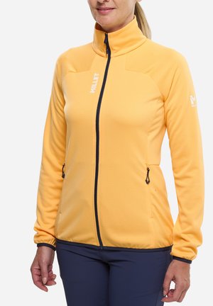 Femme portant une veste jaune à manches longues zippée avec un col montant et une fermeture éclair noire, associée à un pantalon bleu foncé, debout devant un fond blanc.