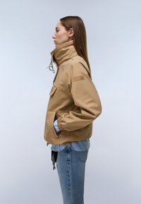 Kobieta z prostymi włosami, ubrana w beżową kurtkę z kieszeniami i elastycznymi mankietami, niebieską koszulę w paski oraz niebieskie dżinsy, stojąca bokiem.