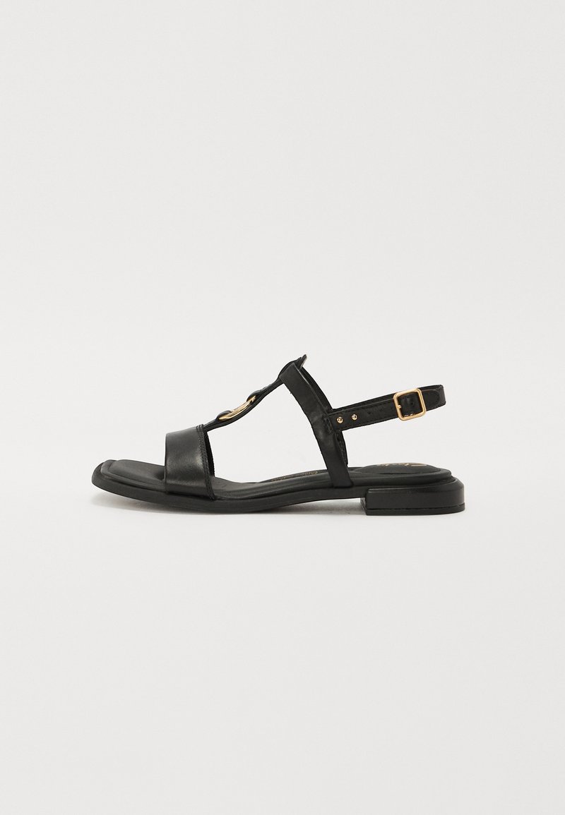 Black leather sandals with a T-strap design, flat sole, gold hardware accent, and adjustable ankle strap. Smooth texture, minimalistic style.