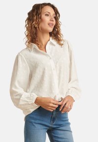 Blouse blanche à manches longues bouffantes, avec un motif en cœur texturé et des poignets à boutons, assortie à un jean en denim bleu.