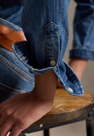 Persoon met opgerolde blauwe denimmouwen die haar arm laat rusten op een houten kruk met metalen frame en zichtbare leren riem.