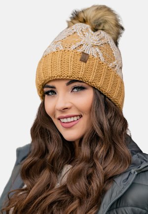 Bonnet en tricot jaune moutarde avec des motifs blancs, présentant un pompon en fausse fourrure sur le dessus et une petite étiquette en cuir sur le côté.