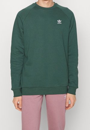 Mies, jolla on päällään tumma vihreä Adidas-huppari ja vaalean mauve Adidaksen verkkarit, seisoo yksinkertaista vaaleaa taustaa vasten.