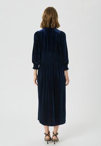 Navyblauw fluwelen jurk met een smocktaille, lange pofmouwen en een middellange snit. Een gladde textuur en zachte val kenmerken het ontwerp.