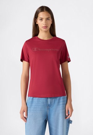 Jeune femme portant un t-shirt rouge Champion avec logo sur la poitrine et la manche, associé à un jean bleu clair sur fond blanc uni.