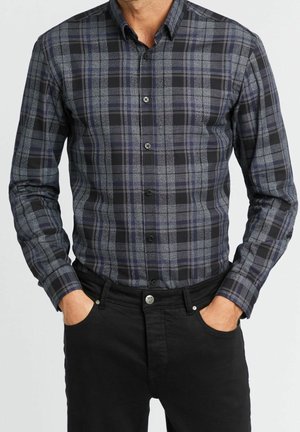 Homme portant une chemise à manches longues à carreaux gris foncé et noir boutonnée, avec un pantalon noir, les mains dans les poches avant.