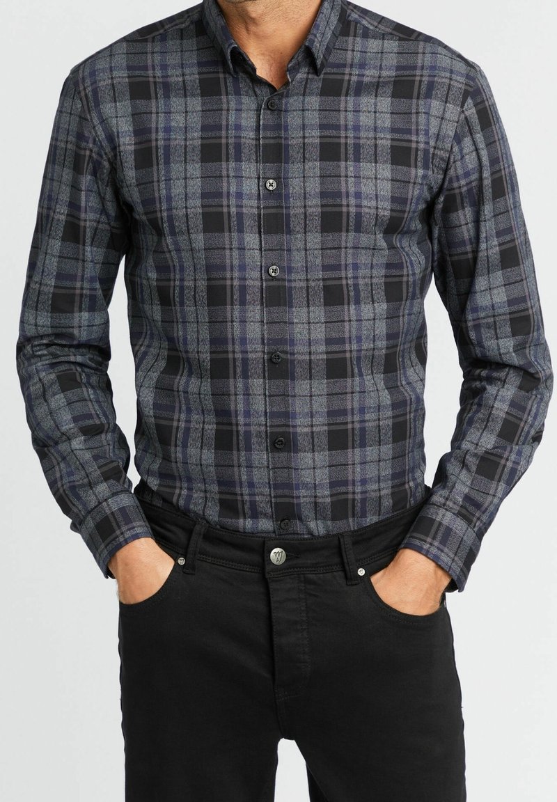 Homme portant une chemise à manches longues à carreaux gris foncé et noir boutonnée, avec un pantalon noir, les mains dans les poches avant.