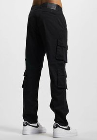 Pantalons cargo noirs avec de multiples poches latérales et un patch logo, offrant une coupe décontractée et un tissu durable, associés à des baskets blanches.