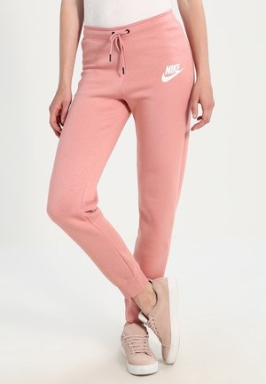 Kobieta w różowych spodniach dresowych Nike ze ściągaczem w pasie oraz jasnoróżowych butach sportowych, stojąca z jedną nogą skrzyżowaną na drugą.