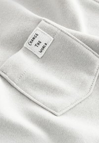 Hellgrauer Stoff mit strukturierter Oberfläche, ausgestattet mit einer rechteckigen Tasche und einem weißen Etikett, auf dem "VERÄNDERE DIE WELT." steht.