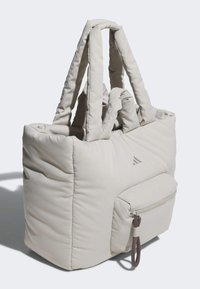 Beige quilted tote bag with padded handles, front zip pocket, and subtle logo. Textured fabric and soft shape enhance ergonomic design.