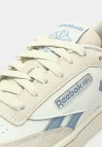 Kraći prikaz bež i bijelih Reebok Classic teniskih obuća s kremastim vezicama i plavim logotipom na strani i jeziku.