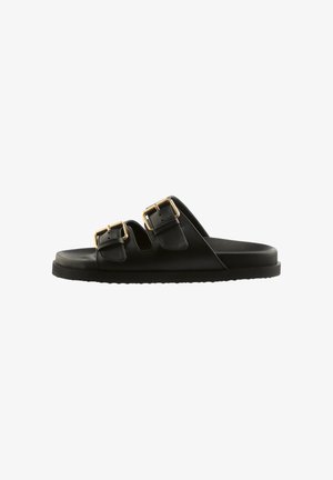 Sandale noire à semelle plate avec deux boucles dorées réglables, vue de côté sur fond blanc.