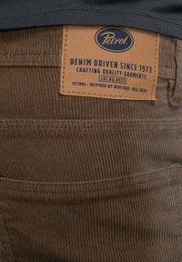 Bruine corduroy stof met verticale ribbels. Een leren label toont het "Petrol" logo en tekst over denim vakmanschap, vastgezet met stiksels.