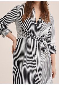 Femme portant une robe boutonnée rayée noire et blanche à manches longues avec un détail de nouage sur le côté, se tenant avec une main sur la hanche.