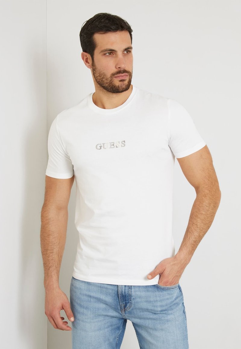 Camiseta blanca de algodón de manga corta con un logo metálico plateado "GUESS" en el pecho, combinada con jeans de mezclilla azul claro.