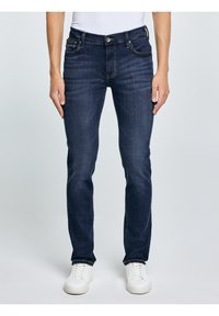 Muž v tmavě modrých džínách slim fit a bílých teniskách, stojící před jednoduchým světle šedým pozadím.