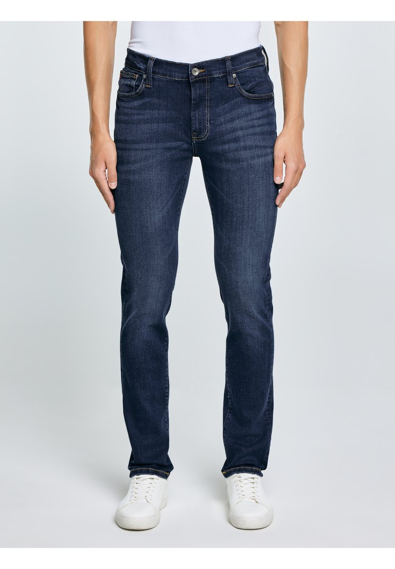 Muž v tmavě modrých džínách slim fit a bílých teniskách, stojící před jednoduchým světle šedým pozadím.