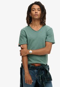 Grøn bomulds T-shirt med V-hals og korte ærmer, parret med blå jeans og en ternet skjorte bundet om taljen. Guldur på håndleddet.