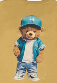 T-shirt présentant un ours en dessin animé portant une casquette de baseball teal et un maillot assorti sur une chemise blanche, avec un jean bleu et des baskets. L'arrière-plan est jaune.