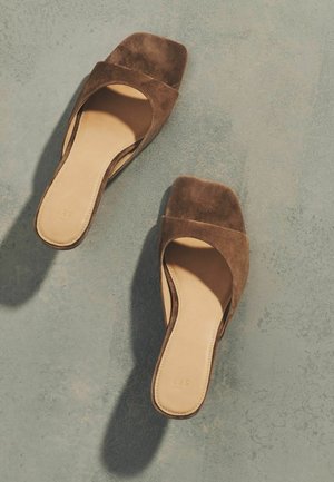 Rudos zomšos atvirų pirštų slidinėjimo sandalų pora su kvadratiniais kulnais, padėta ant tekstūruotos pilkos paviršiaus, metančio šešėlius.