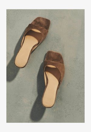 Rudos zomšos atvirų pirštų slidinėjimo sandalų pora su kvadratiniais kulnais, padėta ant tekstūruotos pilkos paviršiaus, metančio šešėlius.