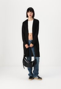 Long black cardigan over a white crop top, paired with blue jeans and black sneakers. Holding a black, round shoulder bag.